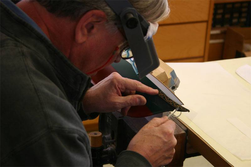A classic model boat builder is rubbing down a custom gold colored foil transfer with a burnishing tool onto the bow of the replica model yacht.