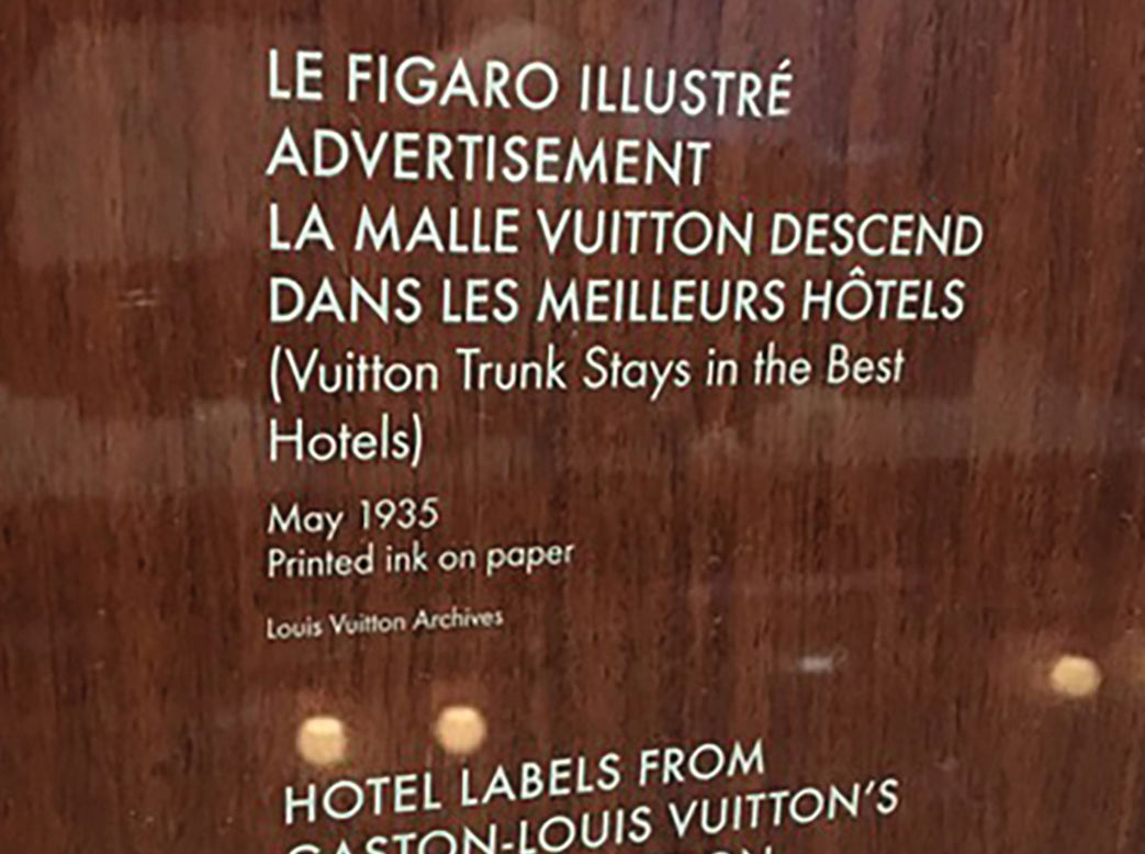 Louis Vuitton - Volez, Voguez, Voyagez exhibit, New York, NY. Custom dry transfer applied to a medium-brown wooden panel, reading, "LE FIGARO ILLUSTRÉ ADVERTISEMENT LA MALLE VUITTON DESCEND DANS LES MEILLEURS HÔTELS (Vuitton Trunk Stays in the Best Hotels) - May 1935 Printed ink on paper - Louis Vuitton Archives". The second one reads, "HOTEL LABELS FROM GASTON-LOUIS VUITTON'S PRIVATE COLLECTION - 20th century Printing on gummed paper - Louis Vuitton Archives".
