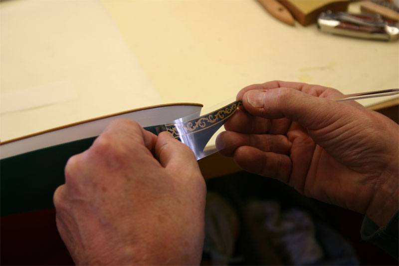 A custom dry transfer is being applied to the bow of the model boat, the gold foil transfer is being peeled from its protective backing strip.